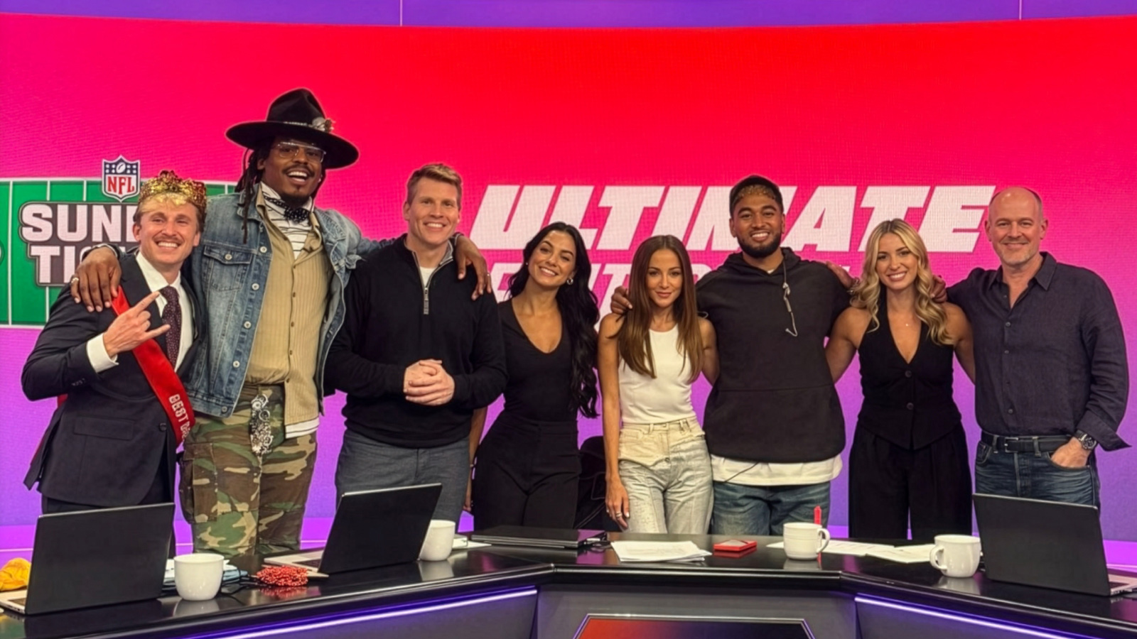 Eight NFL Sunday Ticket hosts smiling together at a purple broadcast studio desk