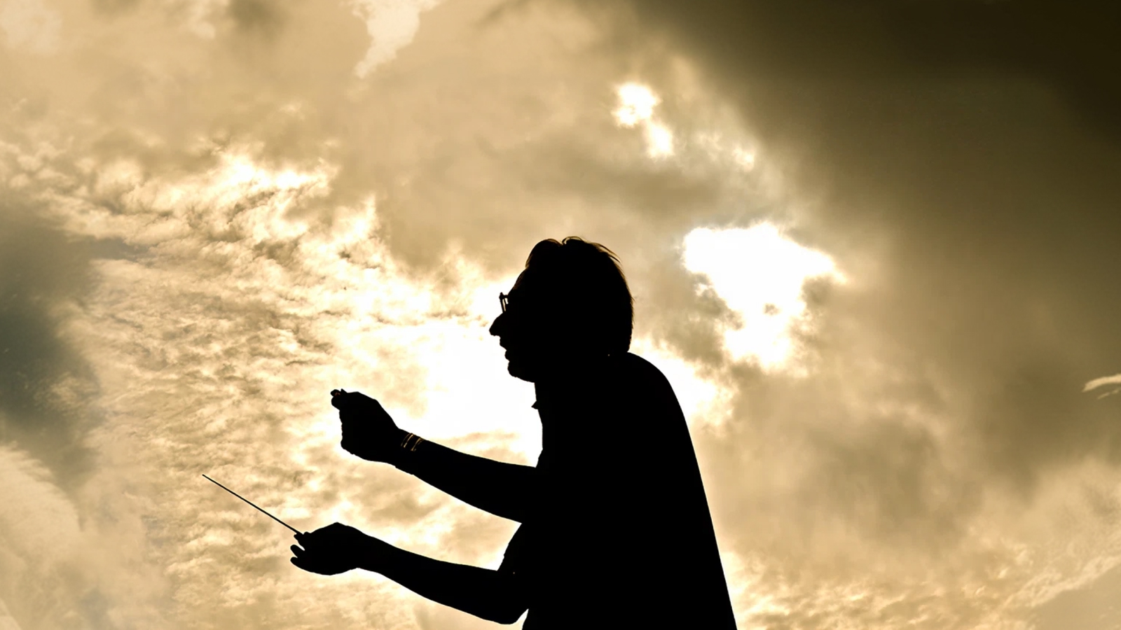 Silhouette of conductor Michael Tilson Thomas raising baton against a golden sky