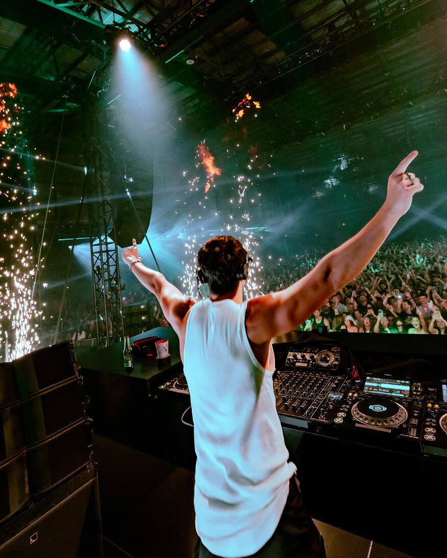 DJ raises arms to a packed crowd amid sparks and stage pyrotechnics during a live show production
