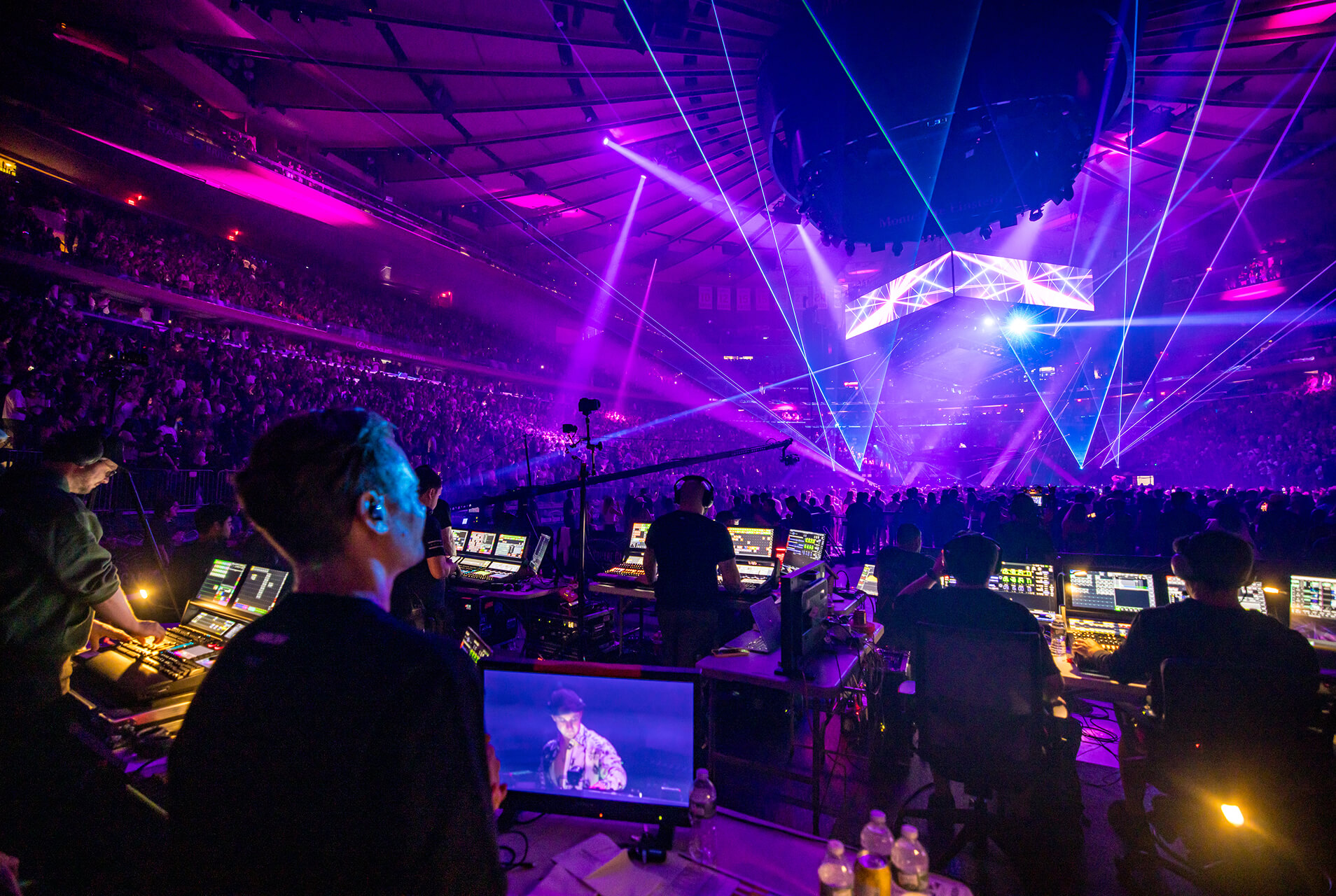 Front-of-house crew at consoles run a purple laser-lit arena concert for a packed crowd