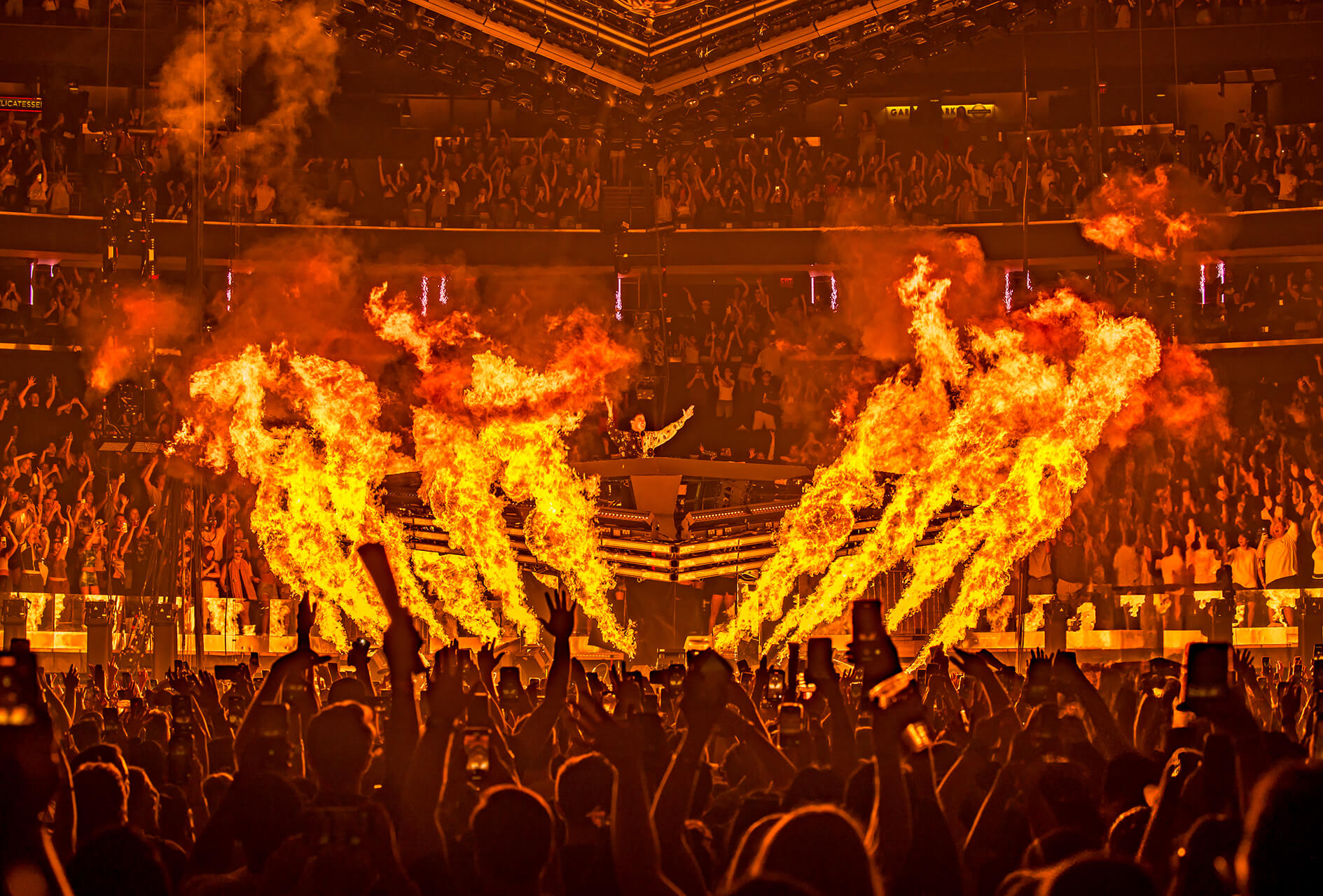 Towering flame jets erupt around a circular DJ stage as fans raise phones at an arena show