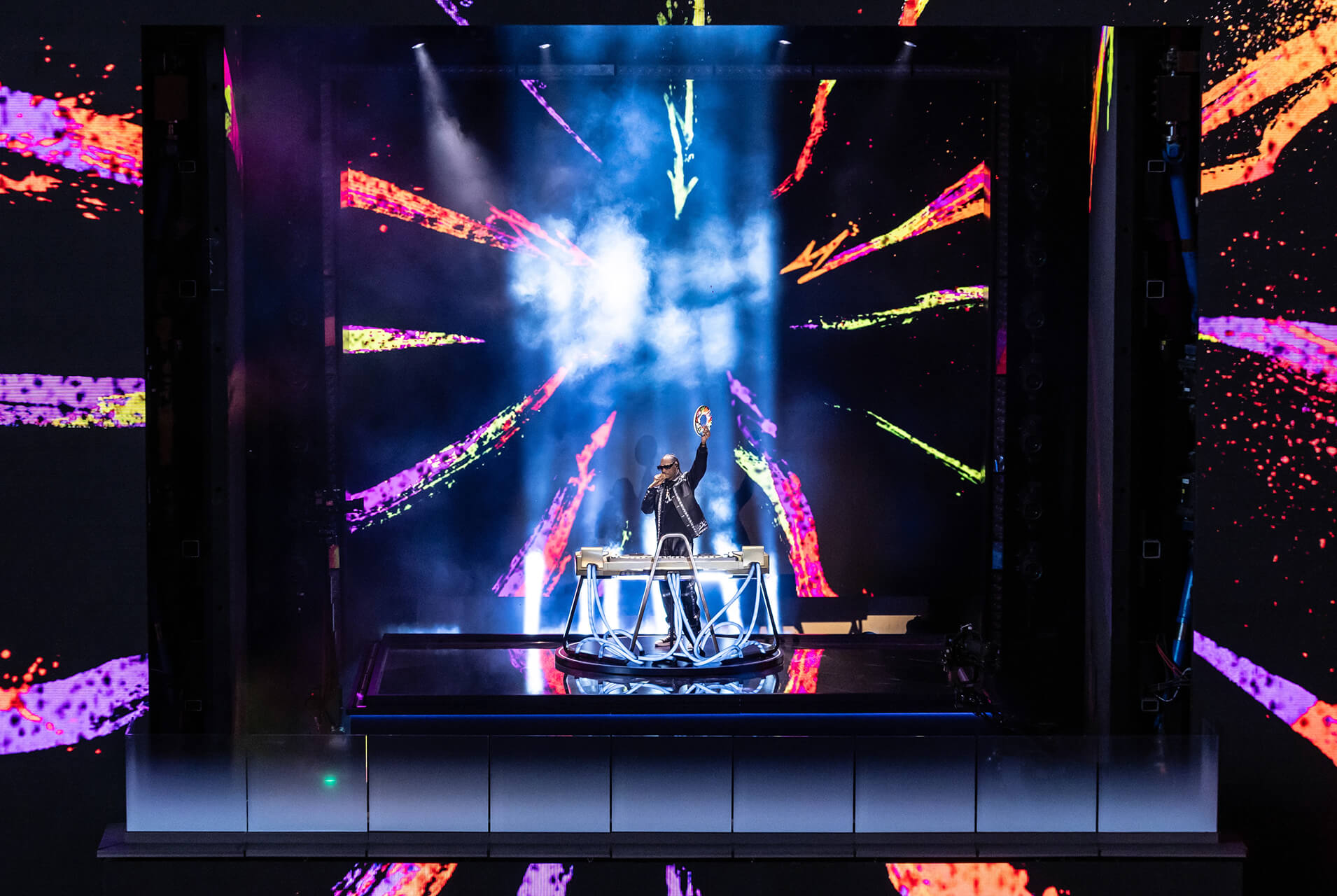 Performer in shades raising a hand onstage before a giant paint-splash LED wall at the Fortnite Chapter 2 Remix launch