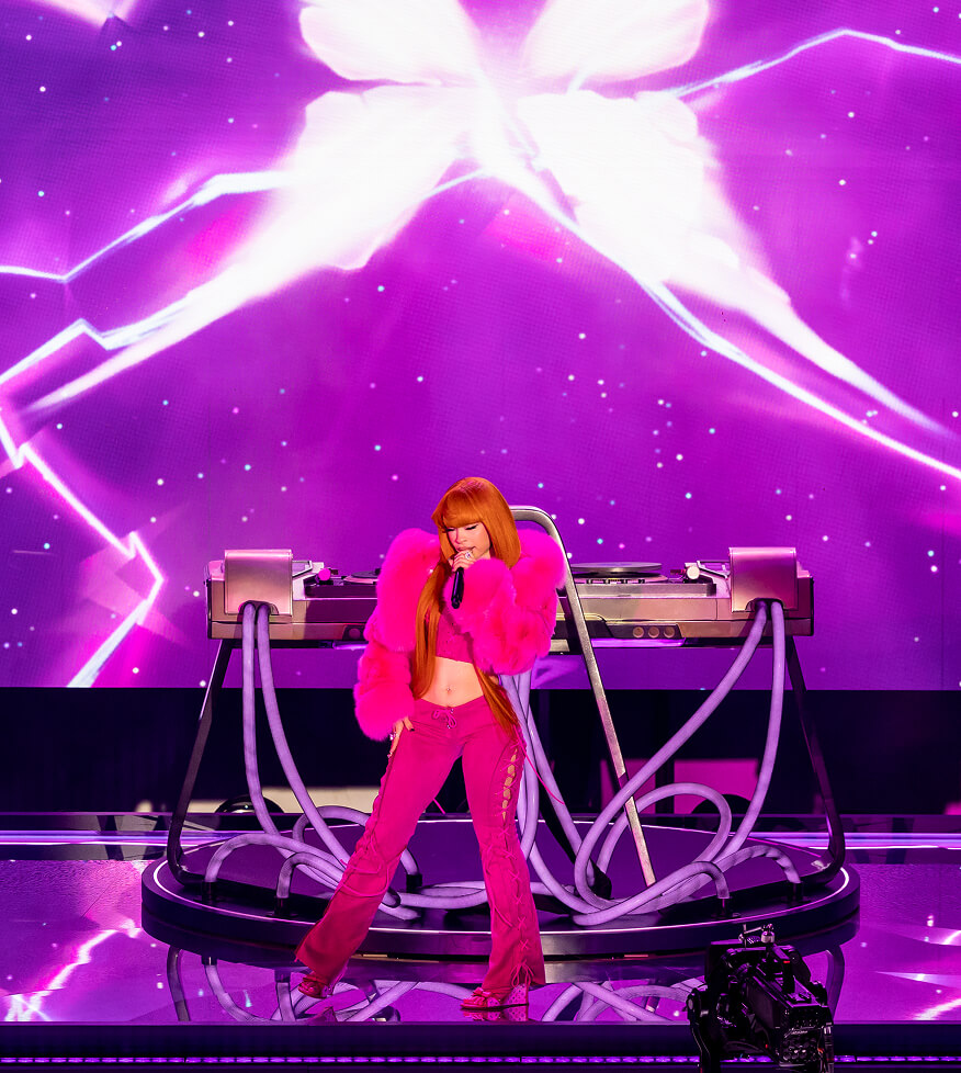 Singer in pink fur performs at a DJ turntable against neon-purple LED visuals, Fortnite Remix launch