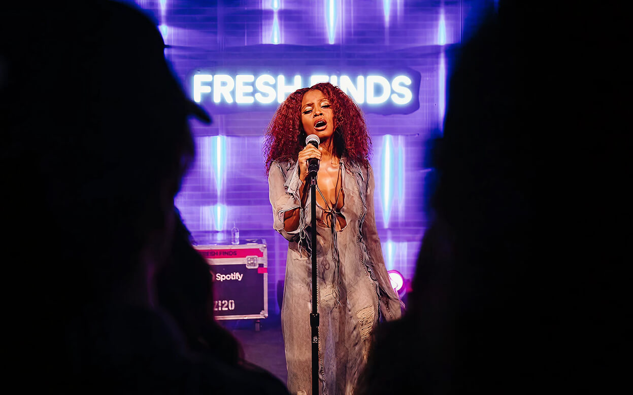 Singer with red curls performs into mic beneath glowing Fresh Finds neon sign at Spotify event