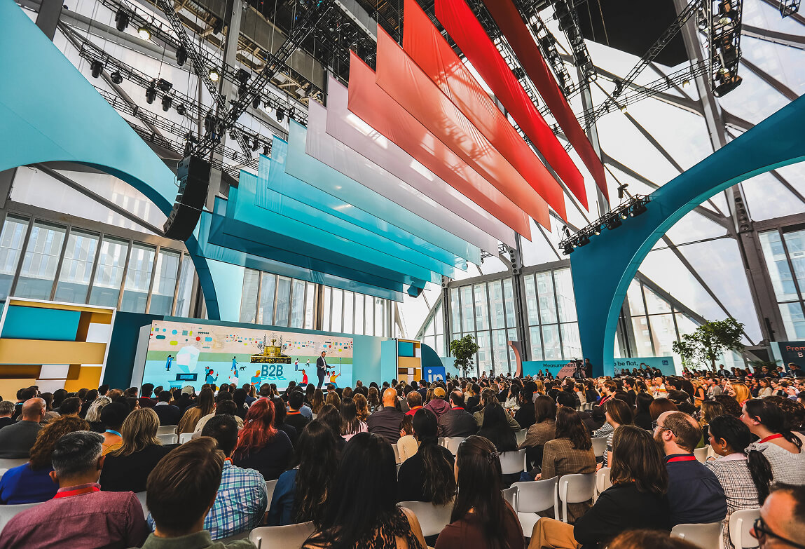 Packed audience faces stage with hanging red and teal banners at LinkedIn B2Believe event