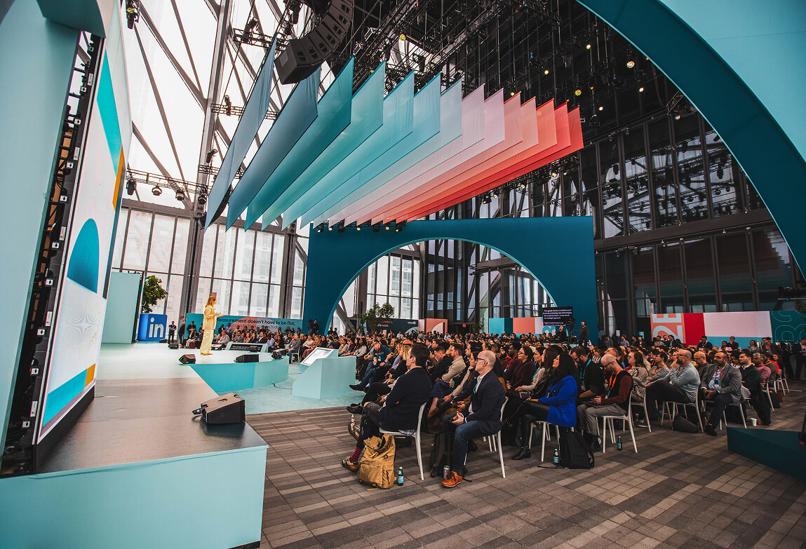 Speaker on stage before seated audience at LinkedIn B2Believe, hanging color panels above
