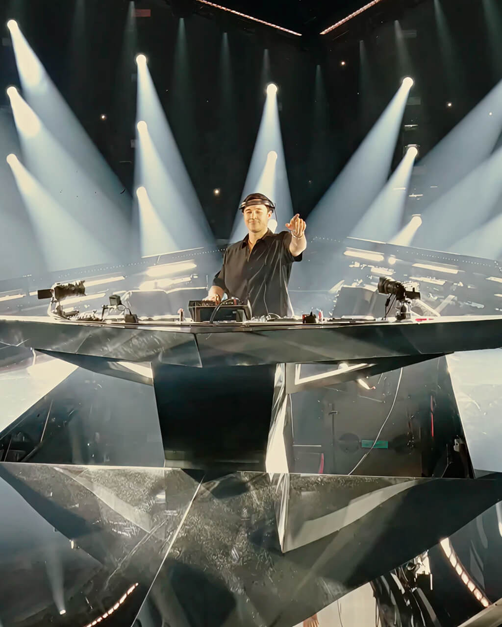 John Summit pointing from his angular DJ booth under white spotlight beams