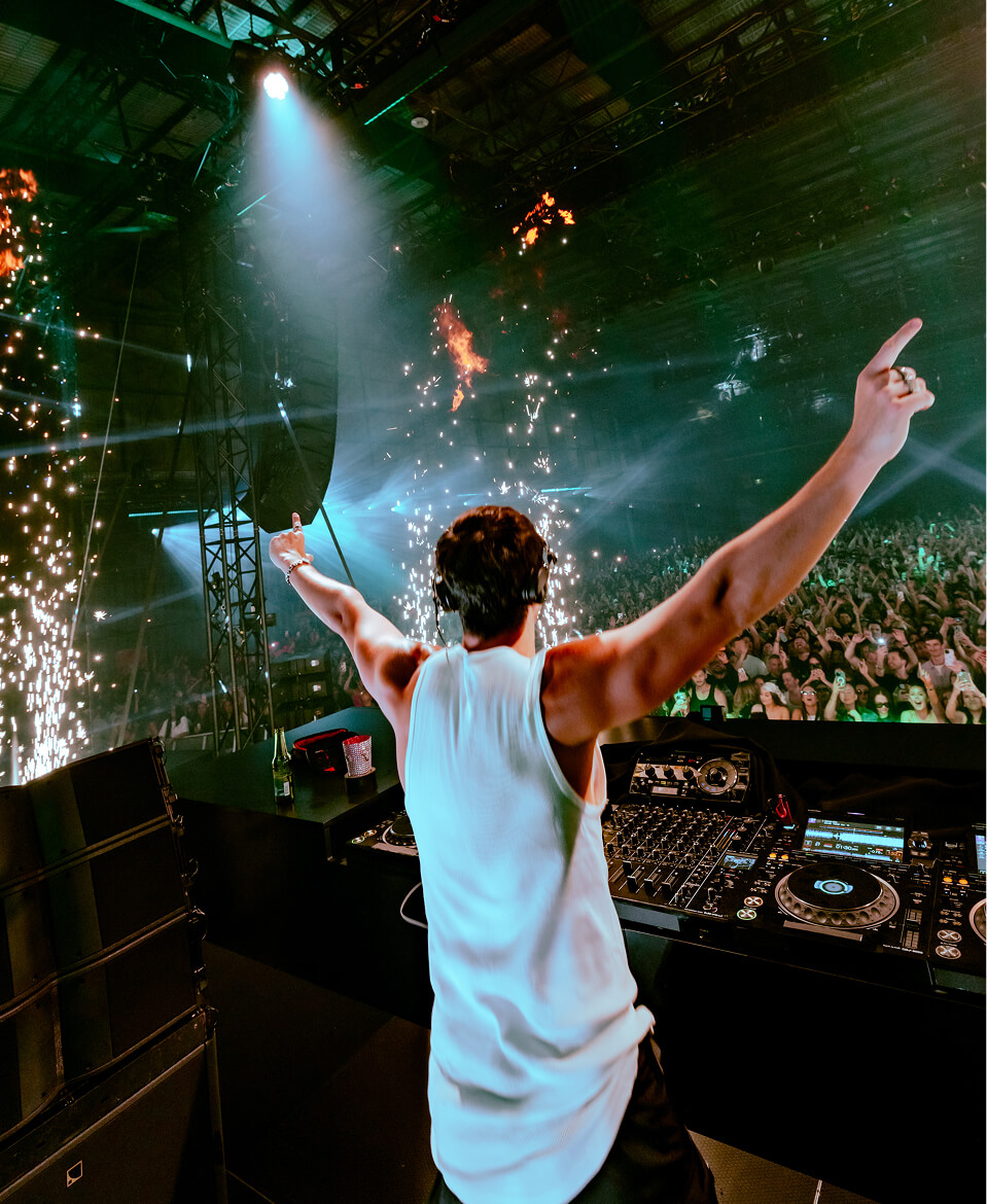 John Summit facing the crowd with arms raised amid sparks and fireworks onstage
