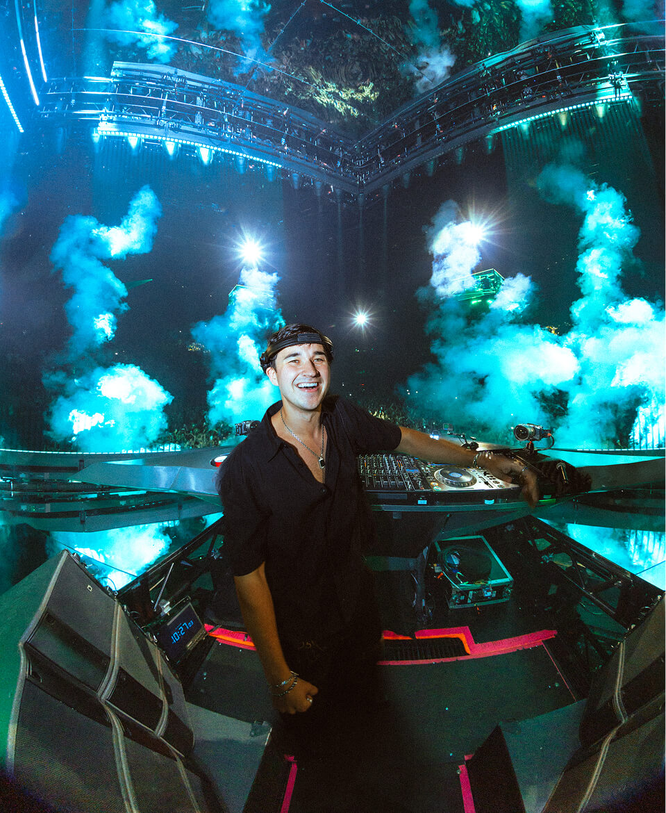 John Summit smiling behind the decks amid blue CO2 smoke plumes at his show