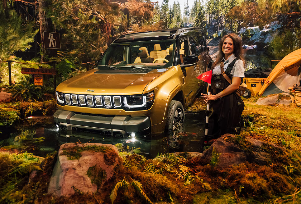 Host poses with gold Jeep Recon crossing a forest stream in the immersive reveal set