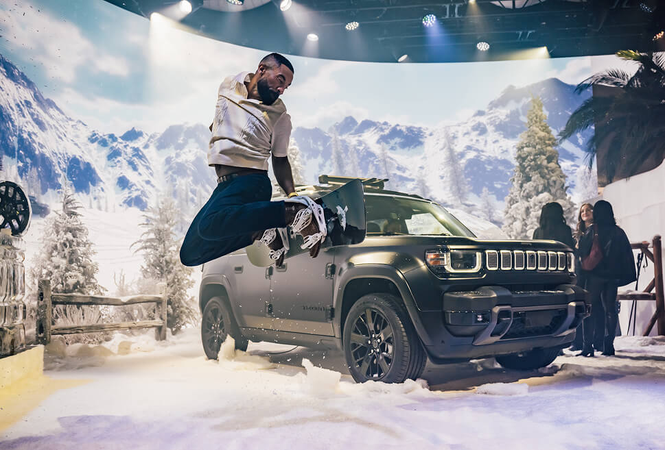Guest leaps mid-air beside a grey Jeep Recon in a snowy mountain set at the reveal