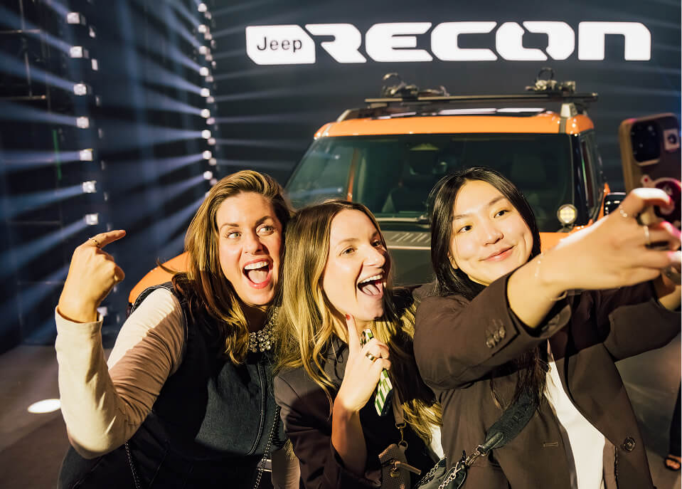 Three friends take a selfie in front of an orange Jeep Recon and glowing logo wall