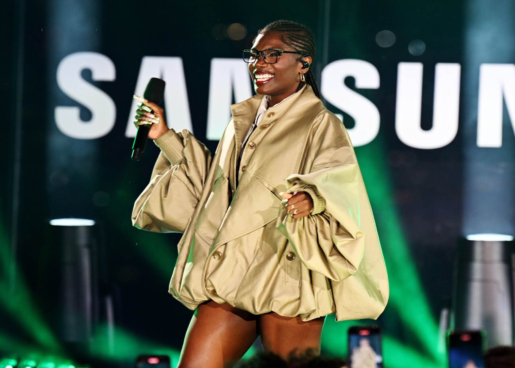 Smiling artist in an oversized khaki jacket performing onstage before a giant Samsung logo