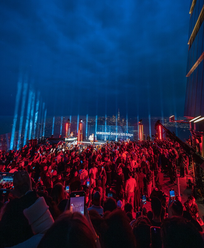Blue searchlight beams over a rooftop crowd facing the Samsung Galaxy S25 Edge stage at night