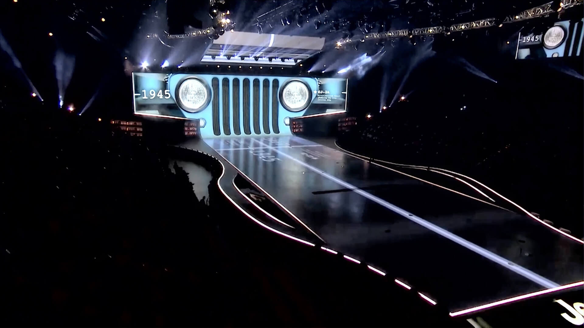 Stage with a giant screen shaped like the front grille of a 1945 Jeep, with a lit runway extending toward the audience.