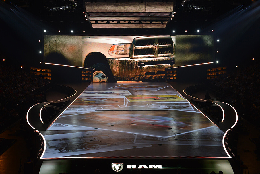 Ram truck reveal stage with a weathered pickup graphic on the screen above a runway lined with vehicle imagery.