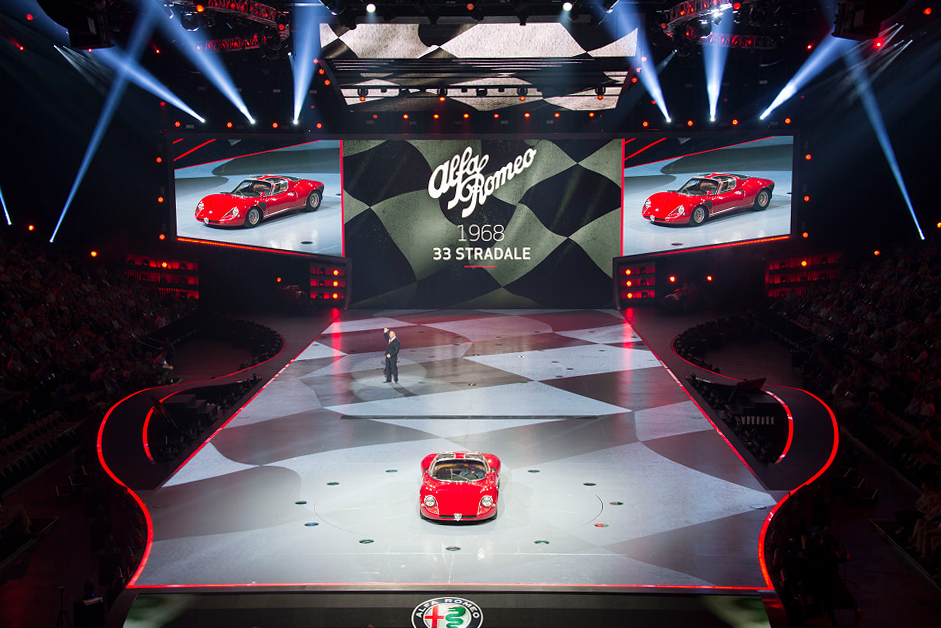 Alfa Romeo stage reveal with a red 33 Stradale on a checkered runway floor, screens showing the car on either side.