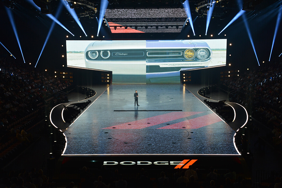 Stage with a giant screen showing a Dodge Challenger split into black-and-white and color halves, with a lit runway extending toward the audience.
