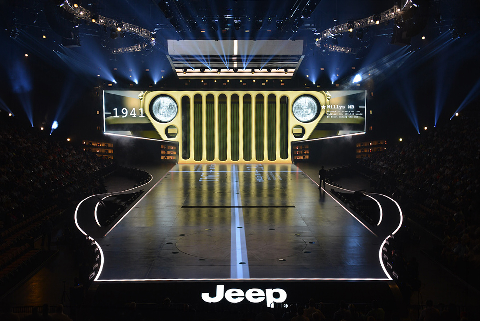 Stage with a giant screen shaped like the front grille of a 1941 Jeep Willys MB, with a lit runway extending toward the audience.