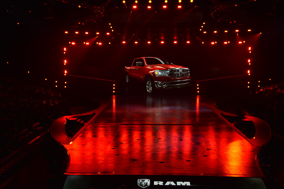 Red Ram pickup truck centered on stage under warm orange lighting, framed by a curved runway.