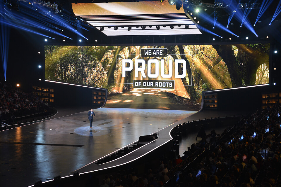 Stage with a forest backdrop reading "We Are Proud of Our Roots," presenter standing at the end of a curved runway.