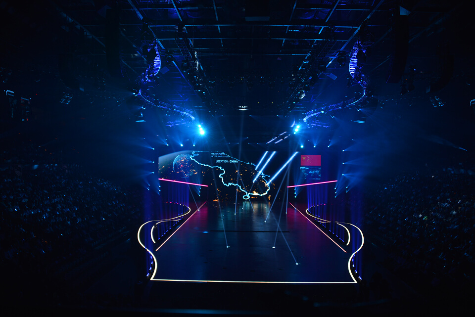 Dark stage with blue light beams and a world map graphic on the center screen, runway extending into the audience.