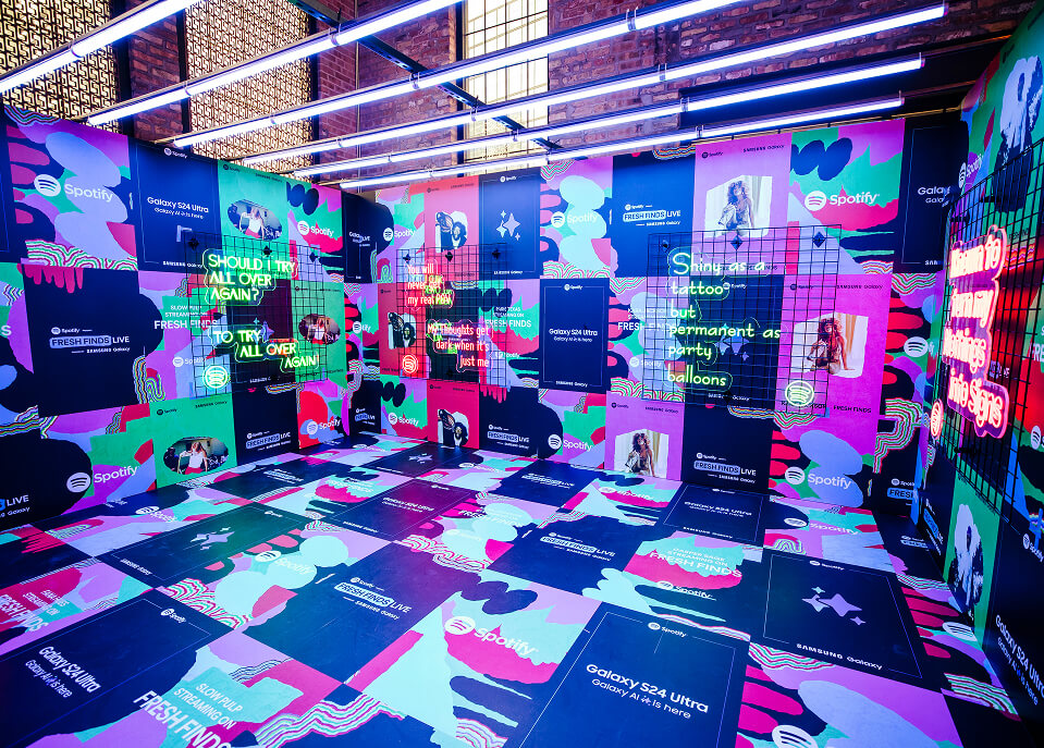 Spotify Fresh Finds Live installation sponsored by Samsung Galaxy S24 Ultra: a room with walls and floor covered in colorful abstract-patterned tiles and logos, with glowing neon phrases mounted on metal grids.